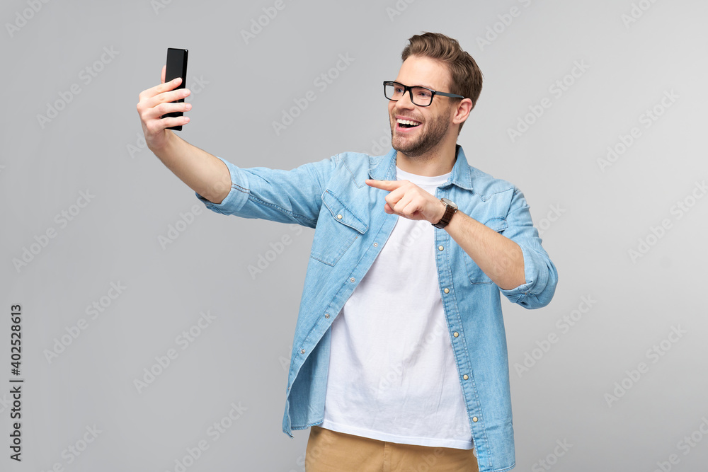 Smiling young man wearing jeans shirt taking selfie photo on smartphone or making video call standing over grey studio background Smiling young man wearing jeans shirt taking selfie photo on smartphone or making video call standing over grey studio background