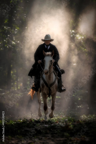 Cowboy is riding a horse in a sunny forest that shines through the big trees.