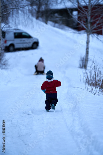 A little boy running after his sisters in the winter. The girls are riding a sleigh or toboggan. 