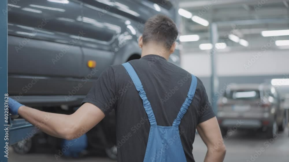 Back view of concentrated Caucasian employee examining vehicle in auto repair shop. Young brunette service man fixing or tuning car indoors. Automobile industry and occupation concept. Back view of concentrated Caucasian employee examining vehicle in auto repair shop. Young brunette service man fixing or tuning car indoors. Automobile industry and occupation concept.