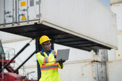 African technician dock worker in protective safety jumpsuit uniform and with hardhat and use digital tablet at cargo container shipping warehouse. transportation import,export logistic industrial
