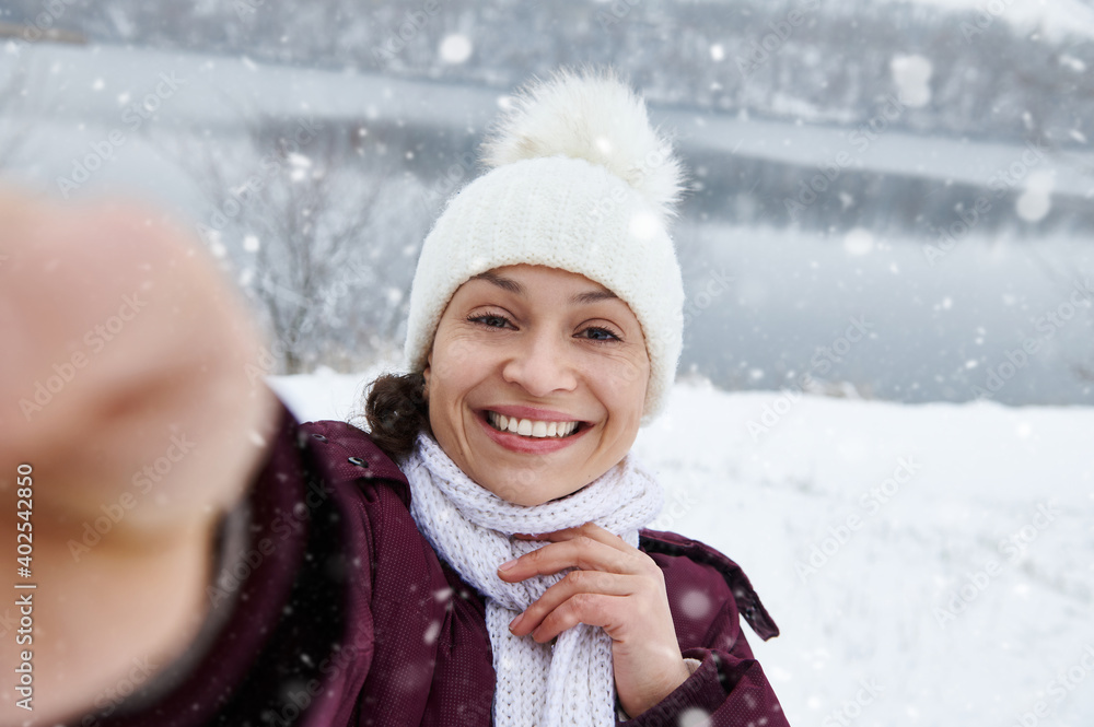 Closeup of a pretty brunette woman smiling at camera while making a selfie enjoying a falling snow on the background of snow covered landscape
