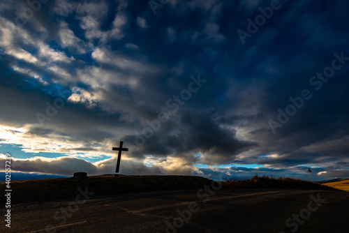 Simple oak wood catholic cross near asphalt road at sunset, dramatic clouds on the sky. 