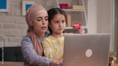 The little girl with her mother in turban is shopping online using a credit cardCute little girl with her mother wearing a headscarf is shopping with a laptop using a credit card. photo