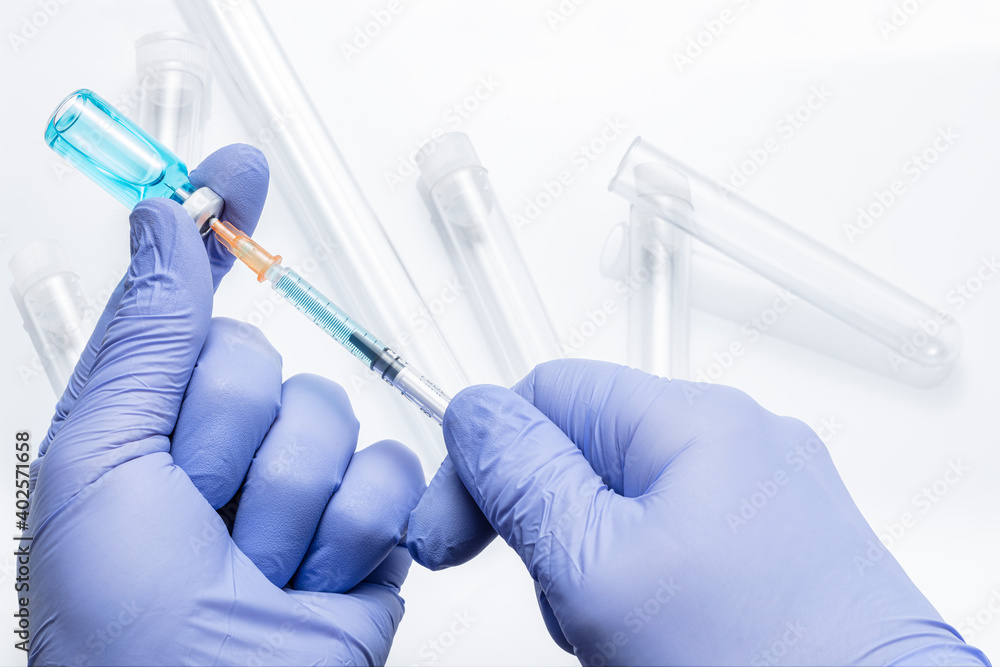 Hands in blue gloves draw medicine from a vial with a syringe on a white background with defocused transparent test tubes Hands in blue gloves draw medicine from a vial with a syringe on a white background with defocused transparent test tubes