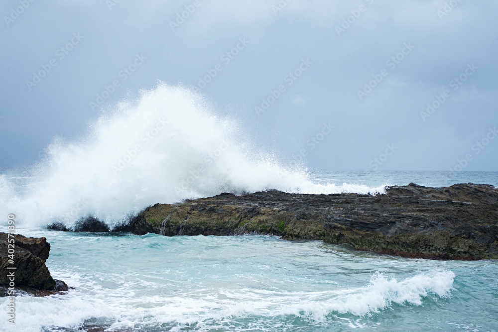 A large wave breaks over rocks sending white spray high into the air. Stormy waves on a gloomy day