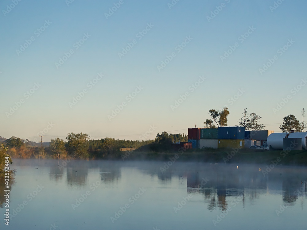 Shipping containers sit piled next to a lake in the early morning as steam rises from the water