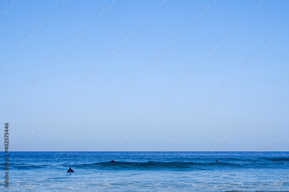 Obraz premium A minimalistic surf line up. Blue skies and blue waters with a small group of surfers sat on thier boards, Cornwall, UK