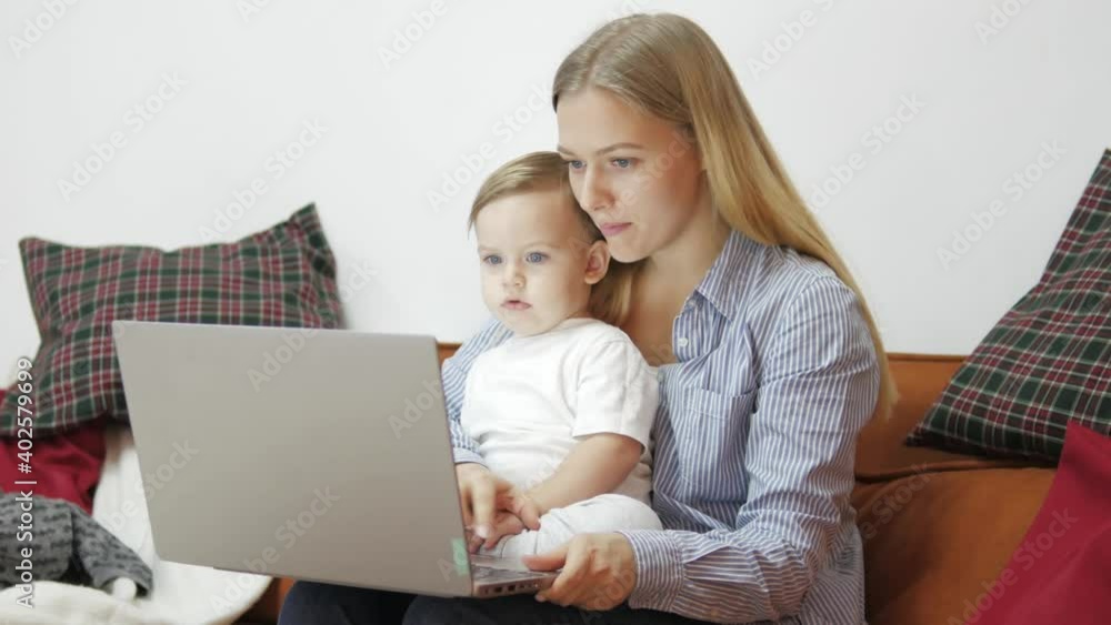 Caucasian Mother multi-tasking, holding baby infant and using computer laptop at home. Candid authentic and real life mom working and parenting sitting on a sofa in the living room Caucasian Mother multi-tasking, holding baby infant and using computer laptop at home. Candid authentic and real life mom working and parenting sitting on a sofa in the living room