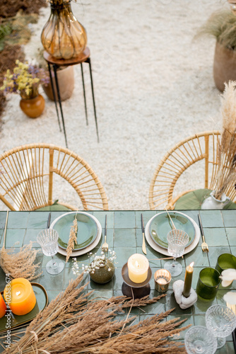 Tablescape of a festivly decorated romantic lunch place in green tones with candles, herbs and flowers in a natural Boho style outdoors. View from above photo