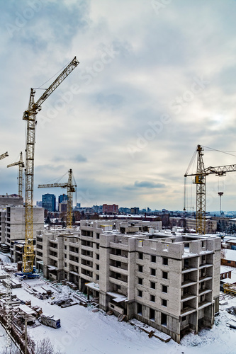 Yellow construction building cranes in winter on the construction site of a large residential modern high-rise building at the background of city and cloudy dramatic sky.