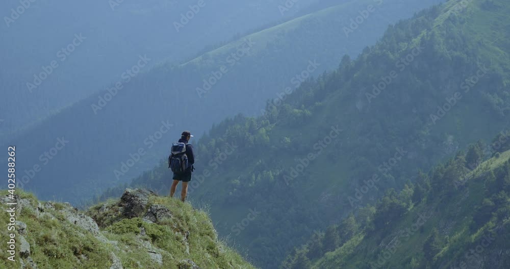 Young man in a cap with a backpack stands on a hill overlooking a valley with mountains and clouds Young man in a cap with a backpack stands on a hill overlooking a valley with mountains and clouds