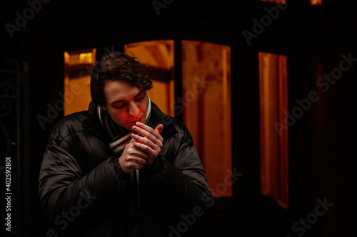 Handsome stylish young hipster man smoking cigarette on the street in winter, looking away. Portrait of beautiful guy dressed casually. Vintage European city, lights on background. Soft focus.