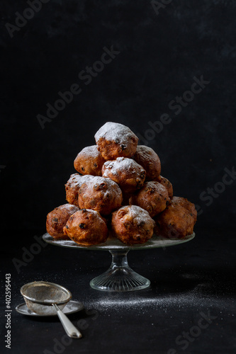 A pile of traditional Dutch oliebollen (deep fried dough balls) with icing sugar on a glass stand on a black background