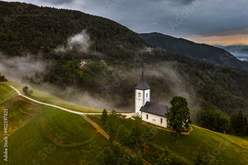 Jamnik, Slovenia - Aerial drone view of a foggy summer sunrise at Jamnik St.Primoz church. The fog gently goes around the small chapel with colorful sky and Julian Alps at background