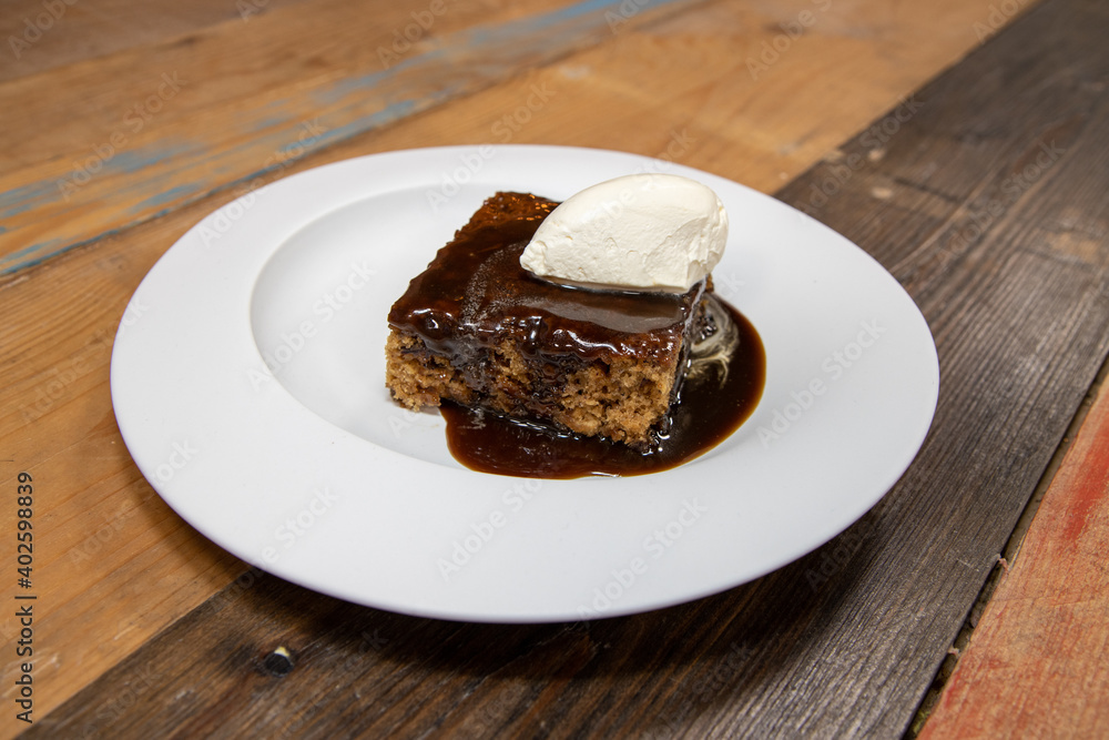 A white plate of delicious sticky toffee pudding chocolate cake with cream and chocolate sauce on a wooden kitchen work top A white plate of delicious sticky toffee pudding chocolate cake with cream and chocolate sauce on a wooden kitchen work top