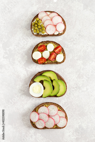 Vegetable sandwiches on slices of dark bread with radish, cucumber, cherry tomatoes, avocado, green peas flat lay on light concrete background
