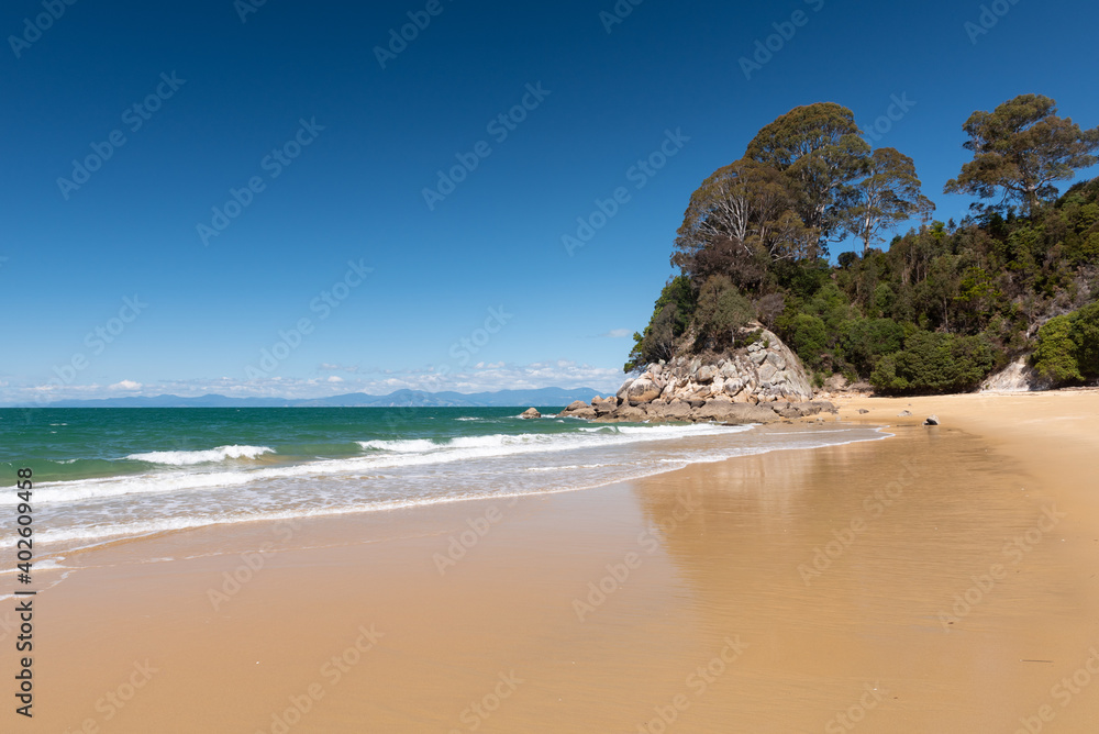 The sandy beach at Breaker Bay, with Kaka Point in the background reflected in the wet sand in the foreground. Kaiteriteri, New Zealand.