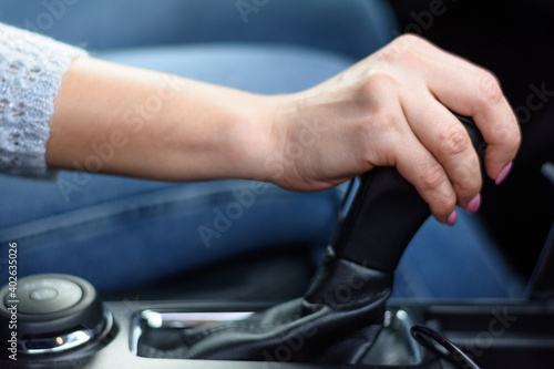 Young woman in jeans and sweater is sitting in the car behind the wheel.. Female driver is driving the car. She is shifting gears in automatic geabox before start moving.