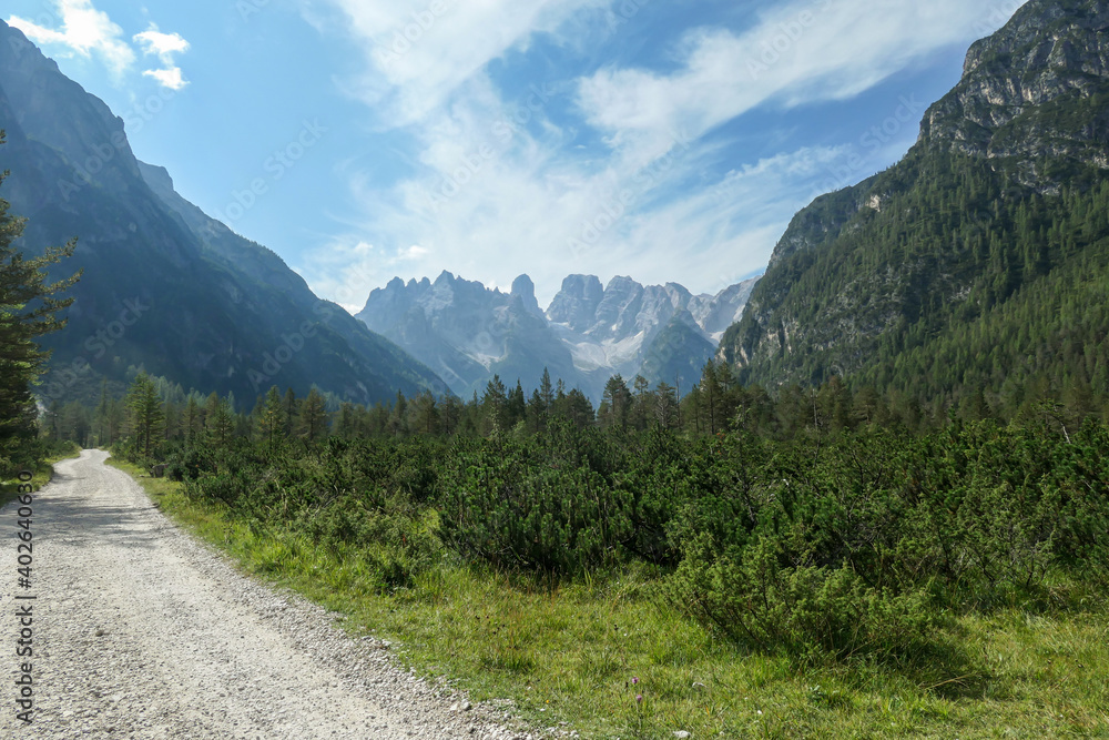 A wide gravelled road in Italian Dolomites from the top of Strudelkopf leading towards the valley. A panoramic view on the high mountains. Sunny day. A few clouds above the peaks. Lush green plateau