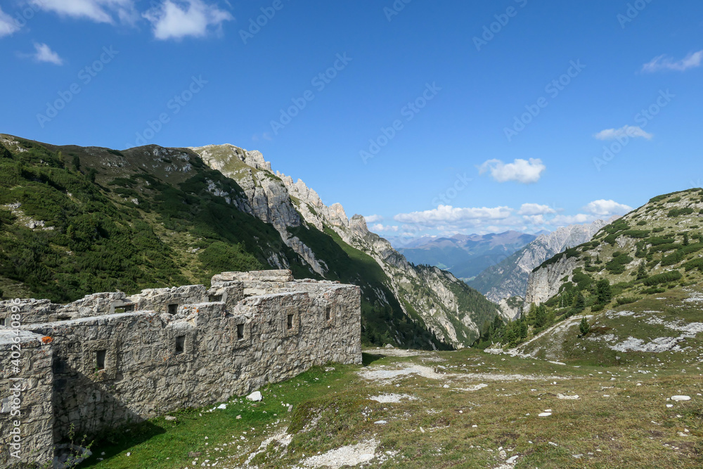 Idyllic view on the high Italian Dolomites from the top of Strudelkopf. A stony construction on the side, resembling a bunker. Sunny day. A few clouds above the high peaks. Lush green plateau around