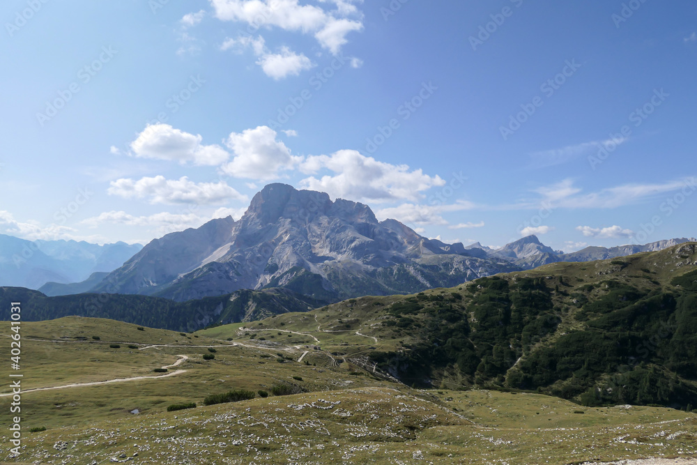 A panoramic view on the high Italian Dolomites from the top of Strudelkopf. There is a wide gravelled path leading to the top. Sunny day. A few clouds above the high peaks. Lush green plateau around
