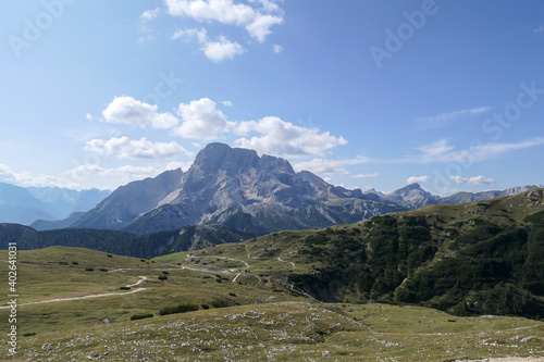 A panoramic view on the high Italian Dolomites from the top of Strudelkopf. There is a wide gravelled path leading to the top. Sunny day. A few clouds above the high peaks. Lush green plateau around