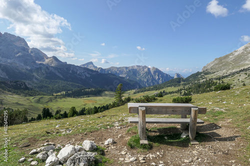 A wooden bench with the panoramic view on the high Italian Dolomites. The are many mountain chains. Sunny day. A few clouds above the high peaks. Lush green plateau around. Meditation and calmness