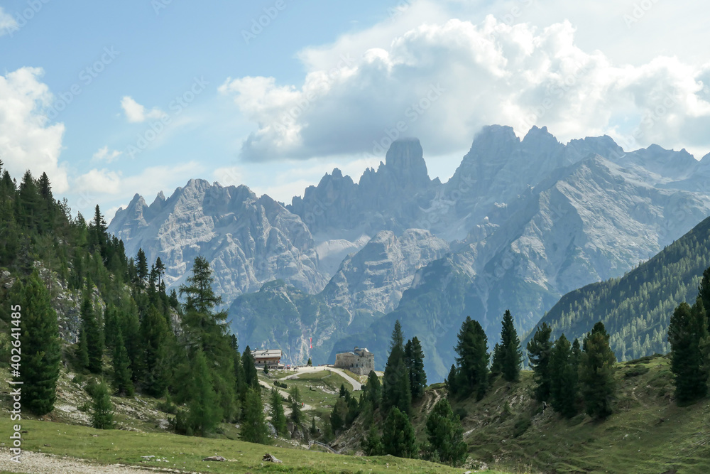 A panoramic view on the high Italian Dolomites, hiding under the clouds. Sunny day. There is a small cottage at the end of a small pathway. Lush green plateau around. Few trees growing in between