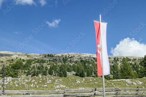 A Tirolian flag on a high pole waving above the high Italian Dolomites. There is a wooden fence behind it. Sunny day. A few clouds above the high peaks. Lush green plateau around with a few trees photo