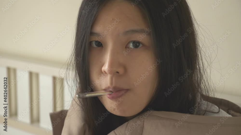 Sick Asian woman sitting in bed, covered in blanket, having cold, shaking, measuring body temperature with thermometer, and looking at the camera. Push in shot, close up, Covid-19 concept Sick Asian woman sitting in bed, covered in blanket, having cold, shaking, measuring body temperature with thermometer, and looking at the camera. Push in shot, close up, Covid-19 concept