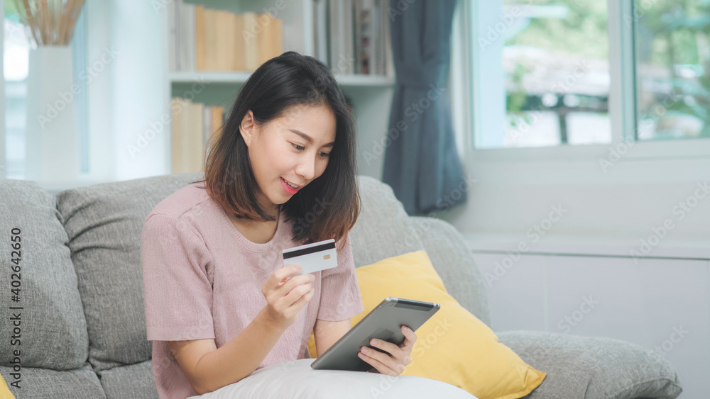 Young smiling Asian woman using tablet buying online shopping by credit card while lying on sofa when relax in living room at home. Lifestyle latin and hispanic ethnicity women at house concept. Young smiling Asian woman using tablet buying online shopping by credit card while lying on sofa when relax in living room at home. Lifestyle latin and hispanic ethnicity women at house concept.