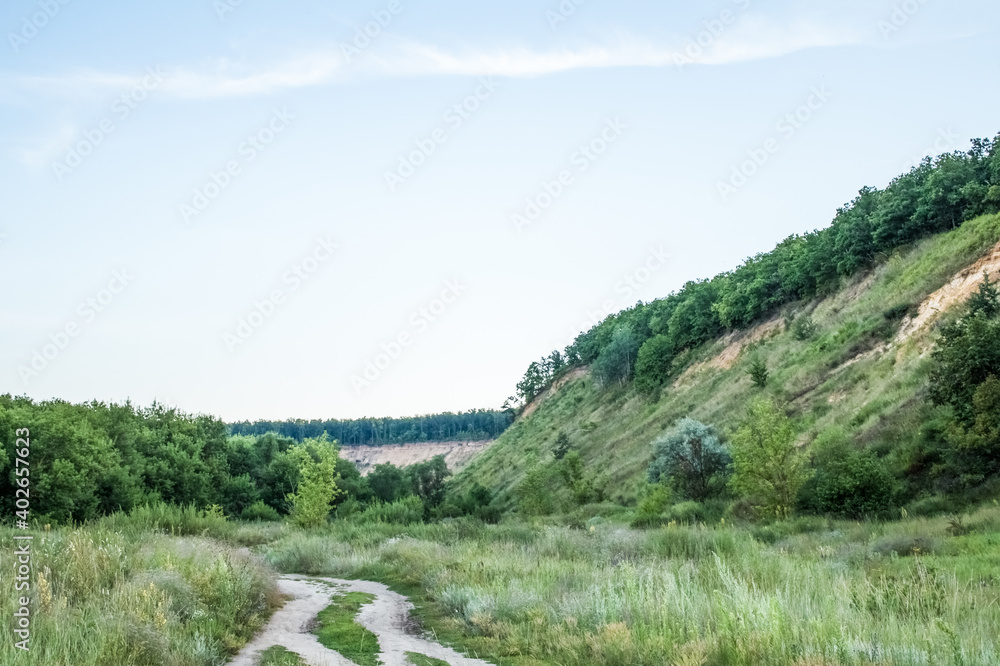 Obraz premium landscaCountry, dirt road goes into the distance across the field. On the sides there are green trees and a sandy mountain covered with plants. Beautiful summer landscape.pe in the mountains