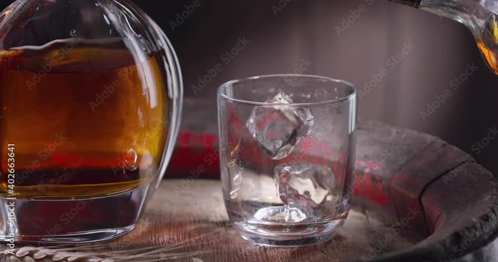 An ice cube slowly falls into a glass, standing on a vintage barrel of whiskey, whiskey from the bottle is immediately poured into the glass. A decanter with a drink stands nearby. An ice cube slowly falls into a glass, standing on a vintage barrel of whiskey, whiskey from the bottle is immediately poured into the glass. A decanter with a drink stands nearby.