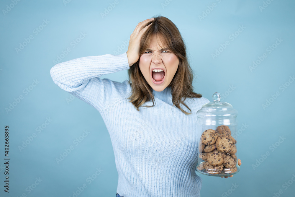 Young beautiful woman holding chocolate chips cookies jar over isolated blue background crazy and scared with hands on head, afraid and surprised of shock Young beautiful woman holding chocolate chips cookies jar over isolated blue background crazy and scared with hands on head, afraid and surprised of shock