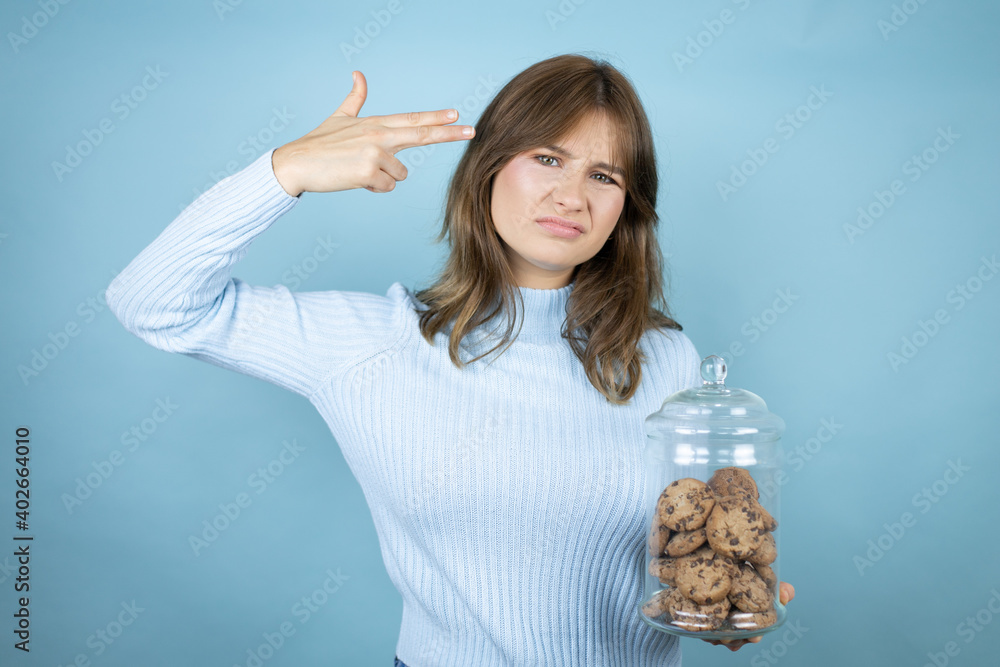 Obraz premium Young beautiful woman holding chocolate chips cookies jar over isolated blue background Shooting and killing oneself pointing hand and fingers to head like gun, suicide gesture.