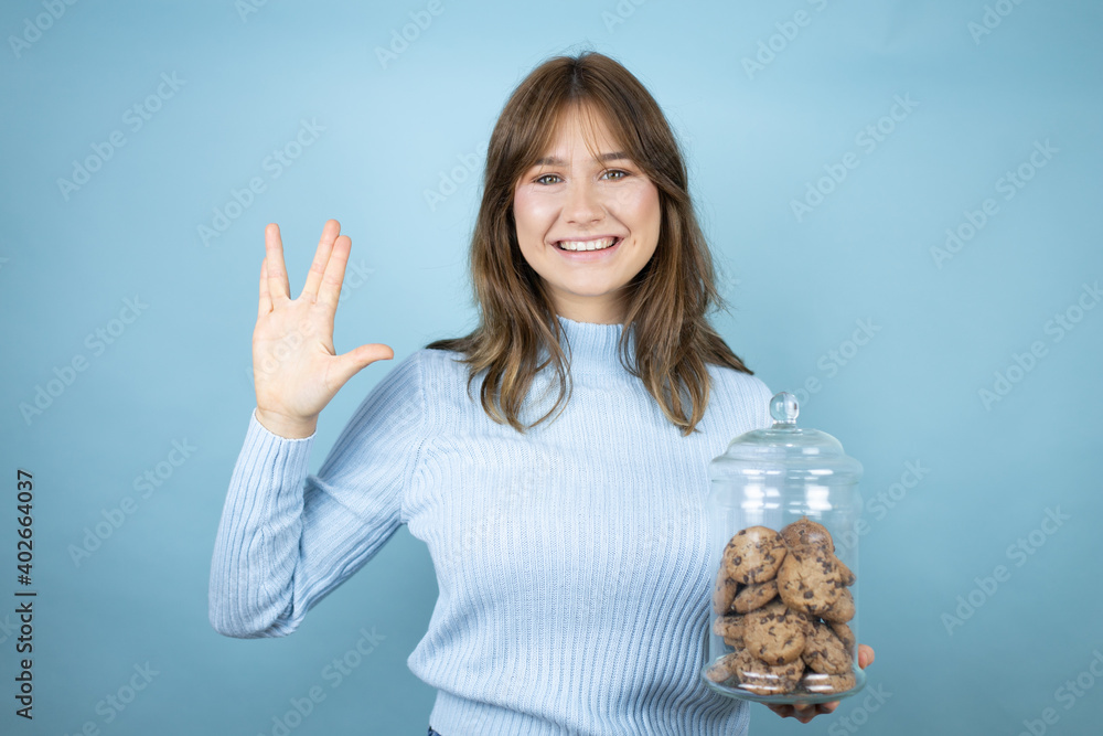 Obraz premium Young beautiful woman holding chocolate chips cookies jar over isolated blue background doing star trek freak symbol