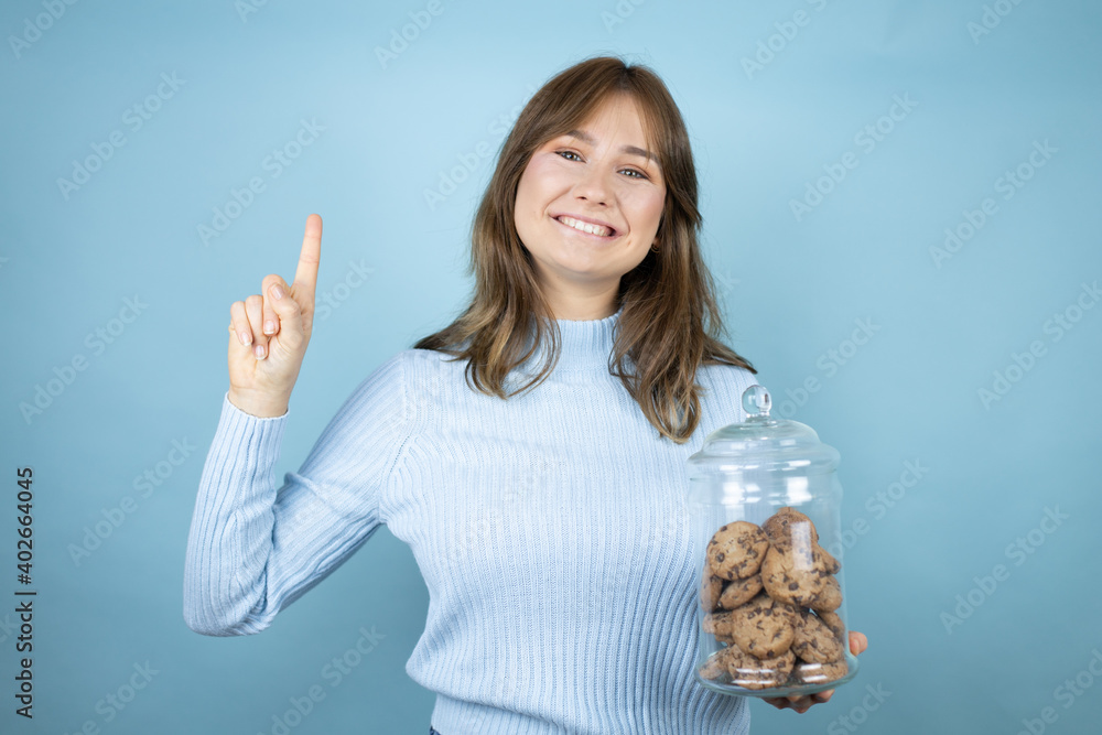 Obraz premium Young beautiful woman holding chocolate chips cookies jar over isolated blue background showing and pointing up with fingers number one while smiling confident and happy