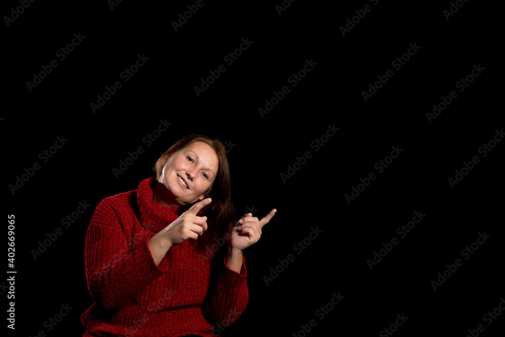 Pretty woman in a red wool sweater pointing fingers to the side on a black studio background. Copy space