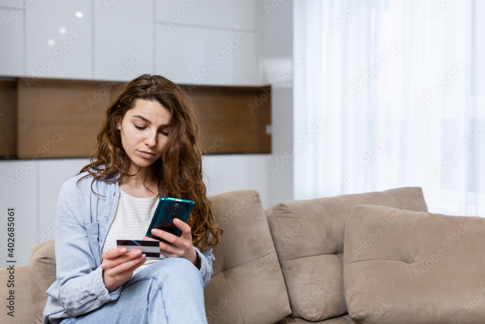 Portrait happy female shopper using instant easy mobile payments making purchase in online store young woman customer holding credit card and smartphone sitting on couch at home Portrait happy female shopper using instant easy mobile payments making purchase in online store young woman customer holding credit card and smartphone sitting on couch at home
