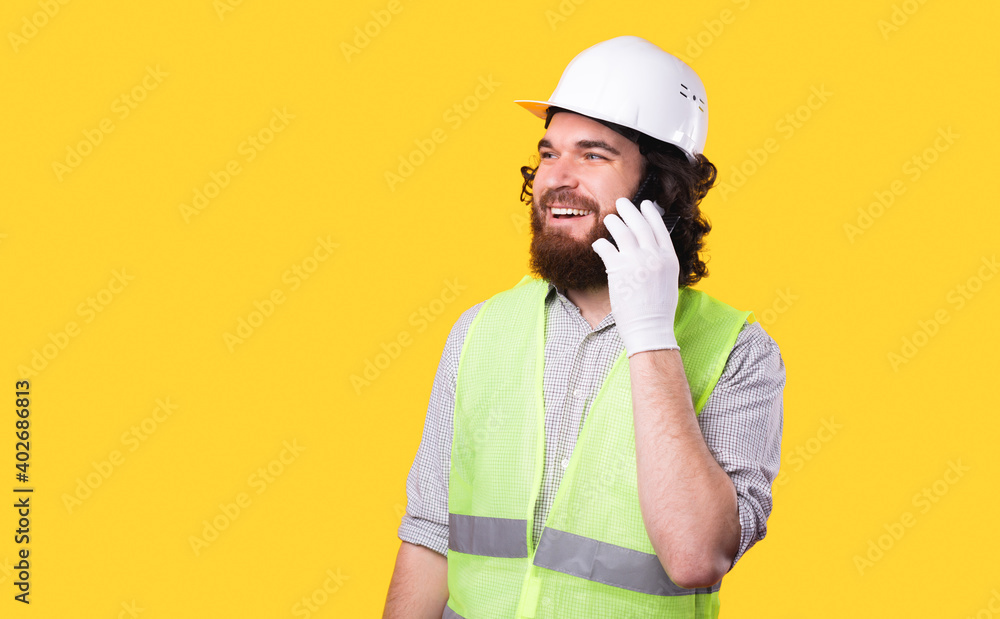 A photo of a cheerful young architect talking on his phone is looking away near a yellow wall . A photo of a cheerful young architect talking on his phone is looking away near a yellow wall .