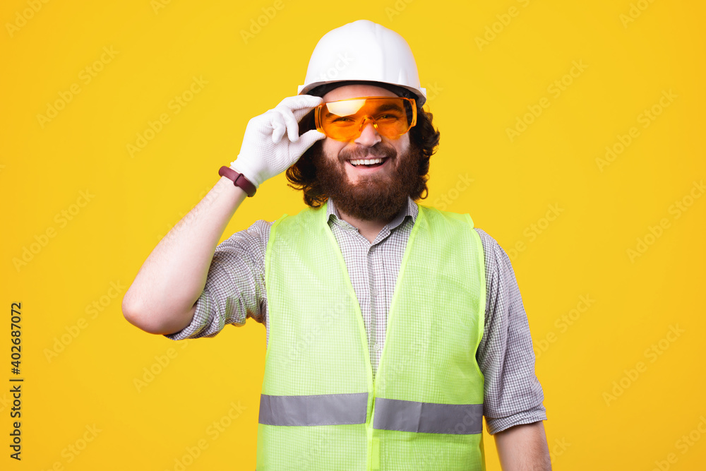 Young cheerful engineer is smiling at the camera and holding a pair of protective glasses is standing near a yellow wall .