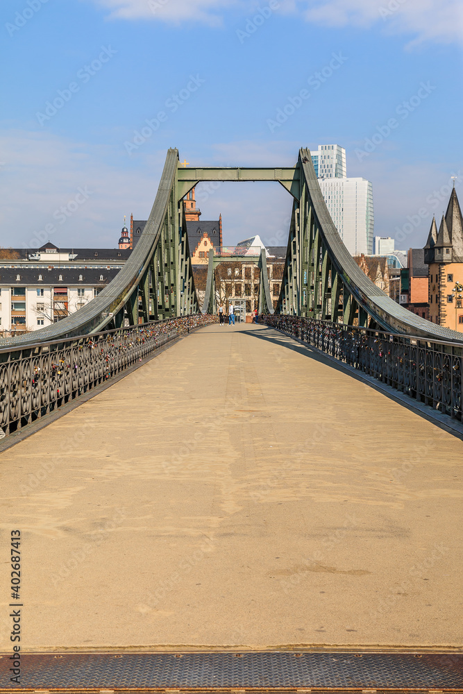 Obraz premium Direct view of the historic pedestrian bridge over the Main in Frankfurt. Houses of the old town in the background with sunshine and a few clouds. Bridge with iron terrain