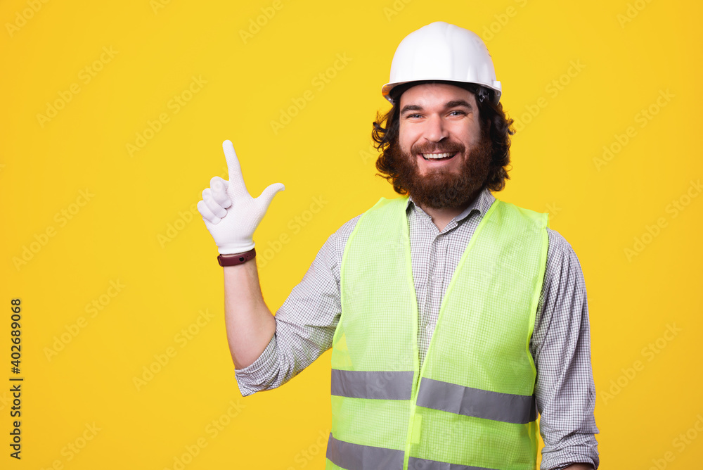 A handsome cheerful young engineer with beard is pointing at a free space smiling at the camera near a yellow wall .
