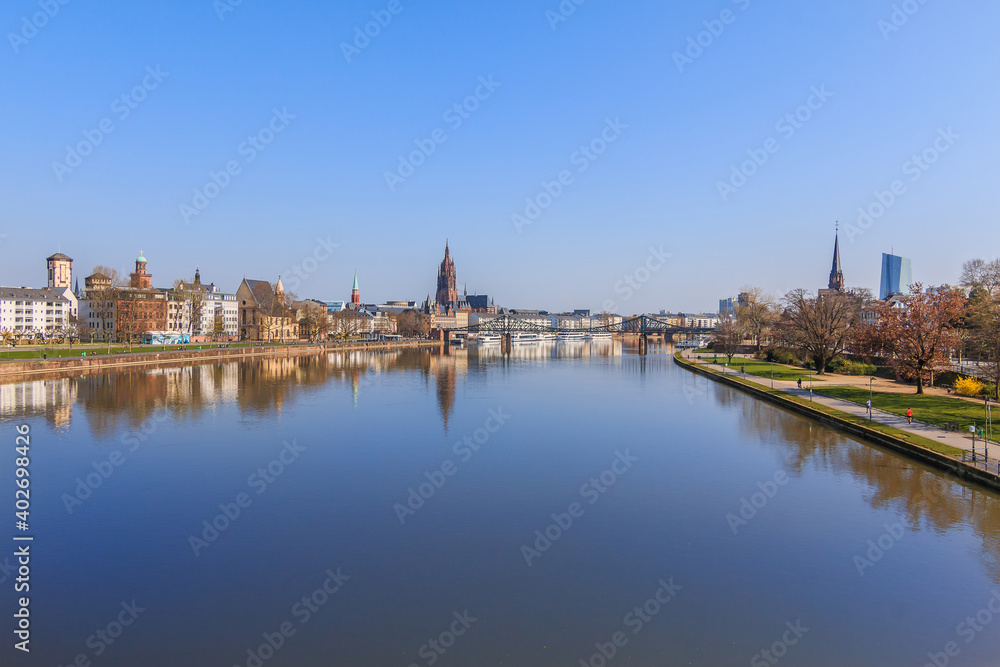 Panorama Frankfurt with the river Main. Park on the banks of the Main with trees and lawns in sunshine and blue sky. Frankfurt Cathedral and buildings of the old town with iron bridge