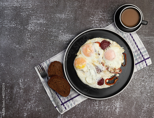 Fried eggs with pepper and sausage and a cup of coffee on on a round plate on a dark background. Top view, flat lay