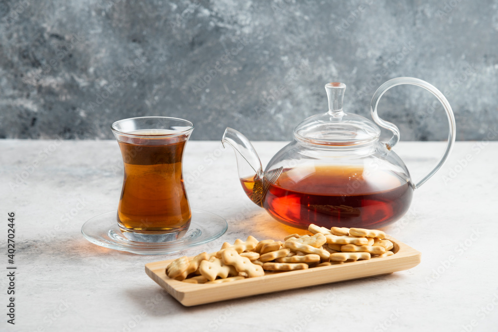 A glass teapot with a cup of tea and a wooden board full of crackers A glass teapot with a cup of tea and a wooden board full of crackers