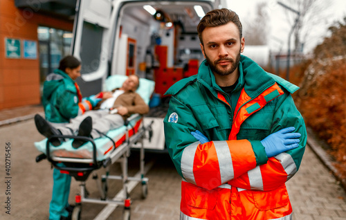 A male paramedic in uniform stands with his arms crossed in front of an ambulance and his colleague standing near a patient's gurney.