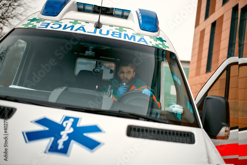 An adult handsome male paramedic is talking on a portable radio while sitting in an ambulance outside a clinic.