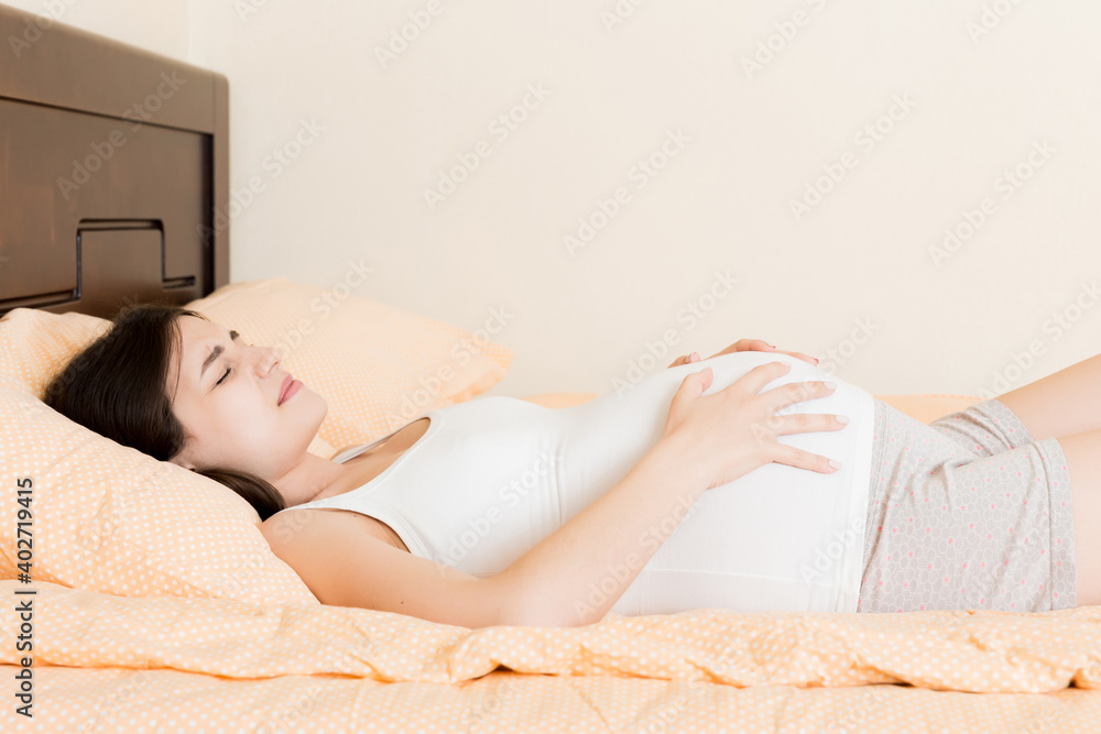 Obraz premium Young pretty pregnant woman, in pain, sitting on bed in bedroom, preparing to give a birth
